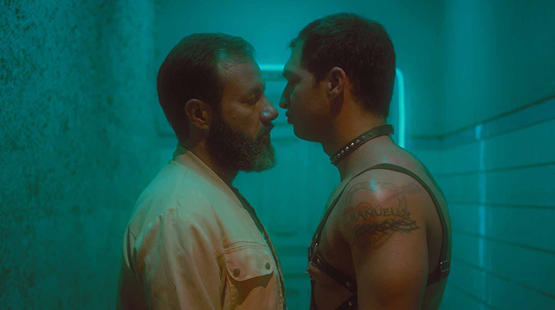 Two men in what appears to be a dim green lit hallway in a bathouse about to kiss