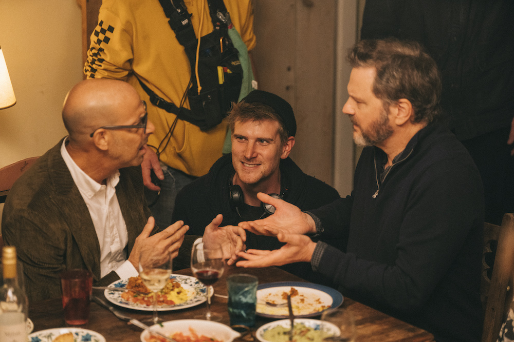 Stanley Tucci, Harry MacQueen and Colin Firth talk at a dinner table in a behind the scenes photo from Supernova film.