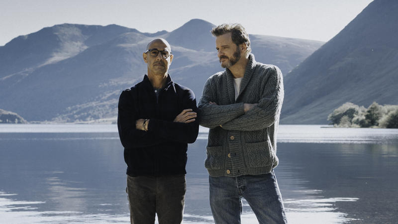 Gay couple Stanley Tucci and Colin Firth stand in front of a beautiful lake and mountain range in Supernova film.